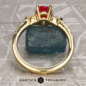 The "Galina" ring in 18k yellow gold with a 1.34-carat Spinel stands upright before a rough blue stone on a textured gray surface, with the Earth's Treasury logo at the bottom.