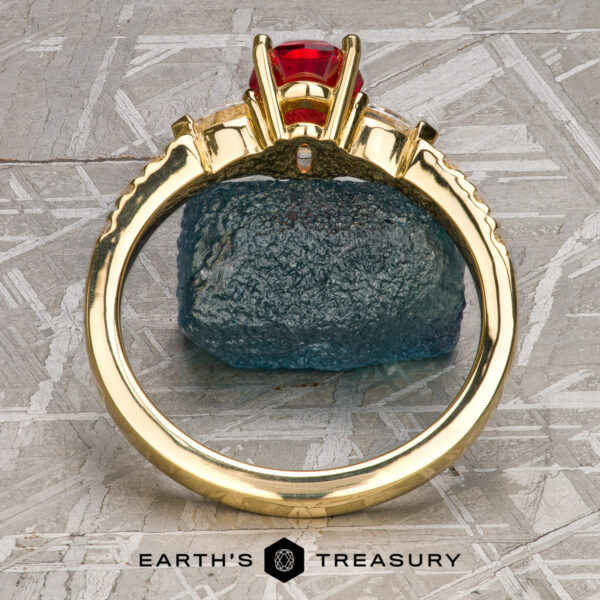 The "Galina" ring in 18k yellow gold with 1.34-carat Spinel The "Galina" ring in 18k yellow gold with a 1.34-carat Spinel stands upright before a rough blue stone on a textured gray surface, with the Earth's Treasury logo at the bottom.