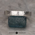 A textured, hammered bandāThe Custom-Fit Straight Bandāis balanced atop a rough, dark gemstone. The band has a rustic, handcrafted appearance, while the gemstone boasts a natural, unpolished texture. In the background lies a neutral, stone-like surface with a subtle geometric pattern. "Earthās Treasury" is elegantly inscribed at the bottom.