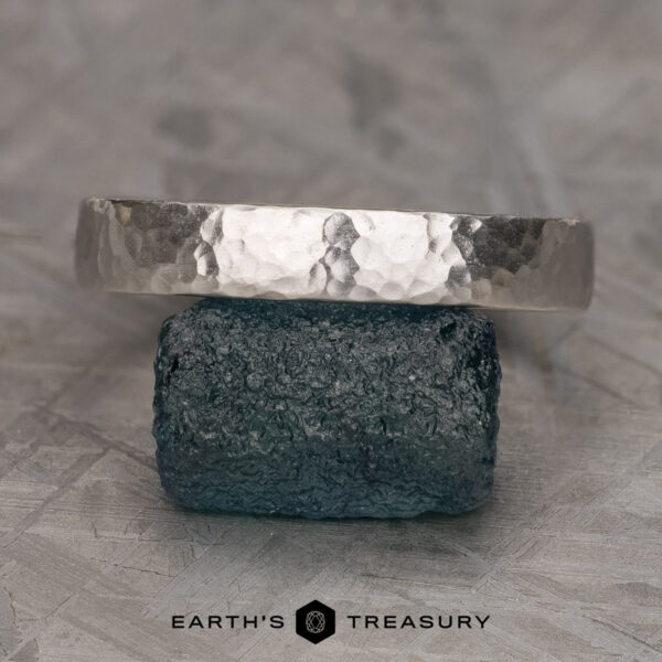 Stacked from 14 images. Method=B (R=1,S=3) A textured, hammered bandāThe Custom-Fit Straight Bandāis balanced atop a rough, dark gemstone. The band has a rustic, handcrafted appearance, while the gemstone boasts a natural, unpolished texture. In the background lies a neutral, stone-like surface with a subtle geometric pattern. "Earthās Treasury" is elegantly inscribed at the bottom.