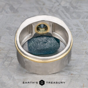 The "Armando" ring, crafted in silver and gold with a 1.57-carat Montana sapphire, stands upright framing a raw blue mineral on a textured gray surface. The "Earth's Treasury" logo appears below.