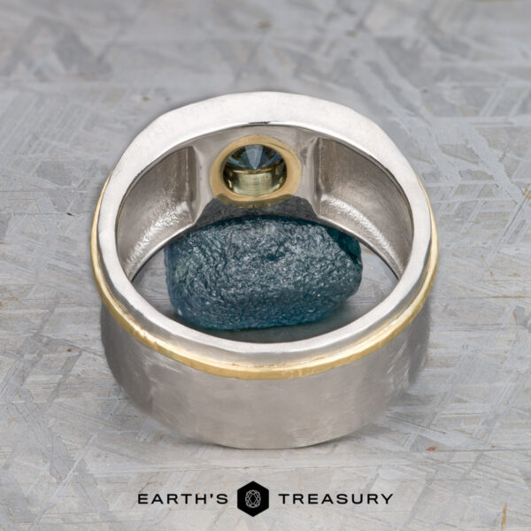 The "Armando" ring with 1.57-carat Montana sapphire The "Armando" ring, crafted in silver and gold with a 1.57-carat Montana sapphire, stands upright framing a raw blue mineral on a textured gray surface. The "Earth's Treasury" logo appears below.