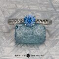 A platinum Engraved "Cathedral" ring with a 0.70-carat Montana sapphire rests on textured blue glass, set against a patterned metallic background. "EARTH'S TREASURY" appears at the bottom.