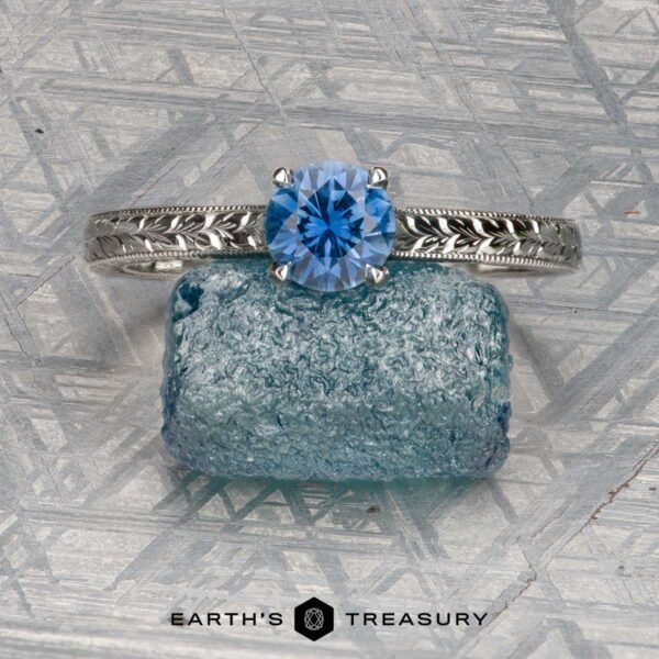 The Engraved "Cathedral" ring in platinum with 0.70-carat Montana sapphire A platinum Engraved "Cathedral" ring with a 0.70-carat Montana sapphire rests on textured blue glass, set against a patterned metallic background. "EARTH'S TREASURY" appears at the bottom.