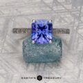 The Engraved "Cathedral" ring in 14K white gold with a 2.75-carat Tanzanite rests on a rough blue crystal, with the Earth's Treasury logo below on a patterned gray surface.