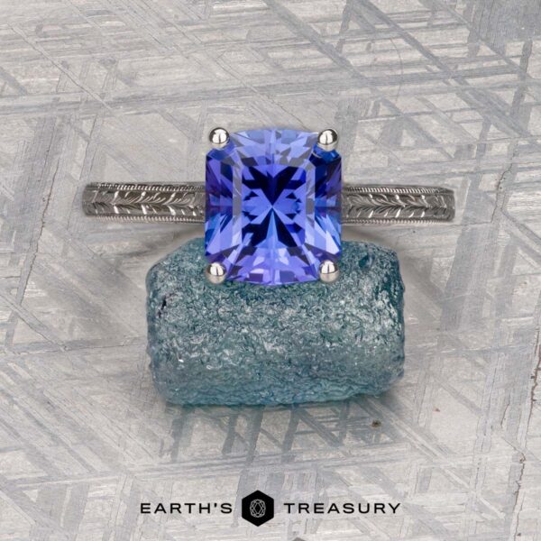 The Engraved "Cathedral" ring in 14K white gold with 2.75-carat Tanzanite The Engraved "Cathedral" ring in 14K white gold with a 2.75-carat Tanzanite rests on a rough blue crystal, with the Earth's Treasury logo below on a patterned gray surface.