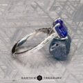 The Engraved "Cathedral" ring in 14K white gold with a 2.75-carat Tanzanite rests on a rough blue crystal, with the Earth's Treasury logo below on a patterned gray surface.