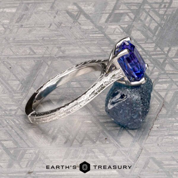 The Engraved "Cathedral" ring in 14K white gold with 2.75-carat Tanzanite The Engraved "Cathedral" ring in 14K white gold with a 2.75-carat Tanzanite rests on a rough blue crystal, with the Earth's Treasury logo below on a patterned gray surface.
