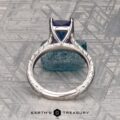 The Engraved "Cathedral" ring in 14K white gold with a 2.75-carat Tanzanite rests on a rough blue crystal, with the Earth's Treasury logo below on a patterned gray surface.