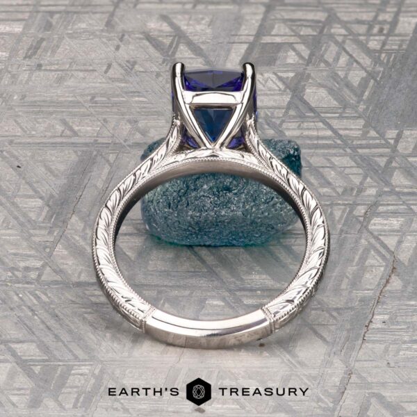 The Engraved "Cathedral" ring in 14K white gold with 2.75-carat Tanzanite The Engraved "Cathedral" ring in 14K white gold with a 2.75-carat Tanzanite rests on a rough blue crystal, with the Earth's Treasury logo below on a patterned gray surface.