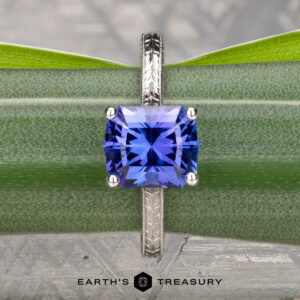 The Engraved "Cathedral" ring in 14K white gold with a 2.75-carat square-cut Tanzanite is displayed vertically against a green plant, with the "Earth's Treasury" logo at the bottom.