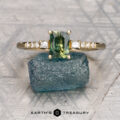 A gold Arsenault, Ben *intl ring featuring a rectangular green gemstone and small diamonds on the band, displayed on a raw blue stone, with the "Earth's Treasury" logo below.