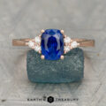 The "Kacie" ring in 14K rose gold features a 1.80-carat cushion-cut Ceylon sapphire and six round diamonds, shown on a textured blue stone with the Earth's Treasury logo below.