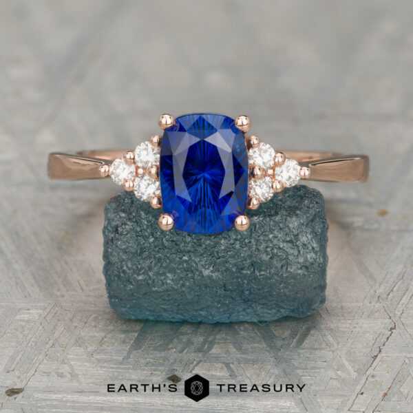 The "Kacie" ring in 14K rose gold with 1.80-carat Ceylon sapphire The "Kacie" ring in 14K rose gold features a 1.80-carat cushion-cut Ceylon sapphire and six round diamonds, shown on a textured blue stone with the Earth's Treasury logo below.
