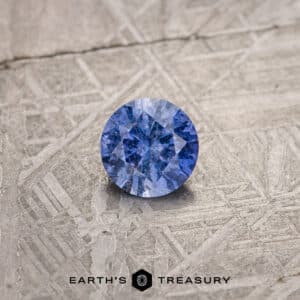 A round, faceted violet purple gemstone is displayed on a textured, metallic surface. The gem has deep violet hues with intricate internal patterns, capturing light and reflecting it with brilliance. The logo and text "EARTH'S TREASURY" are at the bottom of the image.