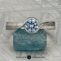 The "Faye" ring in platinum, featuring a 0.92-carat Montana sapphire and diamond-accented solitaire design, is shown on a rough blue crystal against a geometric backdrop with "EARTH'S TREASURY" displayed below.