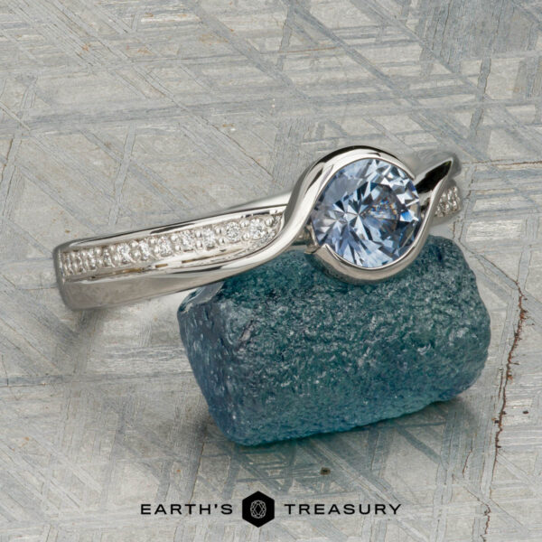 The "Faye" ring in platinum with 0.92-carat Montana sapphire The "Faye" ring in platinum, featuring a 0.92-carat Montana sapphire and diamond-accented solitaire design, is shown on a rough blue crystal against a geometric backdrop with "EARTH'S TREASURY" displayed below.
