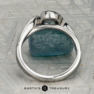 The "Faye" ring in platinum, featuring a 0.92-carat Montana sapphire and diamond-accented solitaire design, is shown on a rough blue crystal against a geometric backdrop with "EARTH'S TREASURY" displayed below.