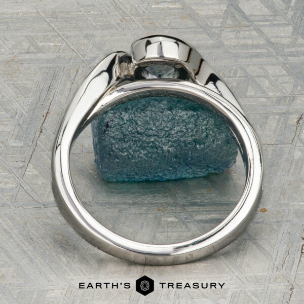 The "Faye" ring in platinum with 0.92-carat Montana sapphire The "Faye" ring in platinum, featuring a 0.92-carat Montana sapphire and diamond-accented solitaire design, is shown on a rough blue crystal against a geometric backdrop with "EARTH'S TREASURY" displayed below.