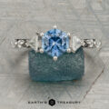 The "Morrigan" ring in platinum, featuring a 1.64-carat Montana sapphire and two clear side stones, is shown upright on a rough blue-green mineral, with the "Earth's Treasury" logo beneath.