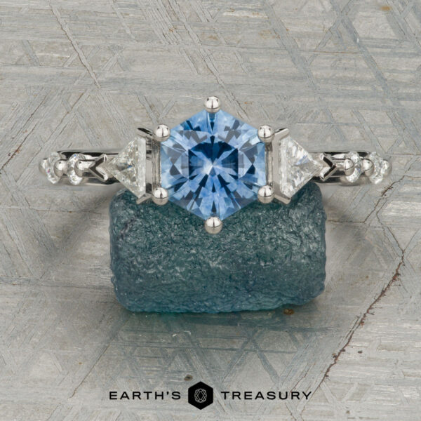 The "Morrigan" ring in platinum with 1.64-carat Montana sapphire The "Morrigan" ring in platinum, featuring a 1.64-carat Montana sapphire and two clear side stones, is shown upright on a rough blue-green mineral, with the "Earth's Treasury" logo beneath.