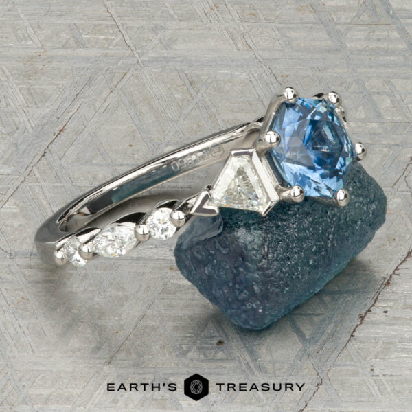 The "Morrigan" ring in platinum with 1.64-carat Montana sapphire The "Morrigan" ring in platinum, featuring a 1.64-carat Montana sapphire and two clear side stones, is shown upright on a rough blue-green mineral, with the "Earth's Treasury" logo beneath.