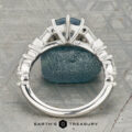 The "Morrigan" ring in platinum, featuring a 1.64-carat Montana sapphire and two clear side stones, is shown upright on a rough blue-green mineral, with the "Earth's Treasury" logo beneath.