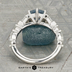The "Morrigan" ring in platinum, featuring a 1.64-carat Montana sapphire and two clear side stones, is shown upright on a rough blue-green mineral, with the "Earth's Treasury" logo beneath.