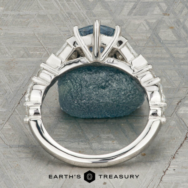 The "Morrigan" ring in platinum with 1.64-carat Montana sapphire The "Morrigan" ring in platinum, featuring a 1.64-carat Montana sapphire and two clear side stones, is shown upright on a rough blue-green mineral, with the "Earth's Treasury" logo beneath.
