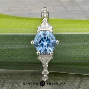 The "Morrigan" platinum ring, featuring a 1.64-carat hexagonal Montana sapphire and two triangular clear stones, is displayed on a green leaf.