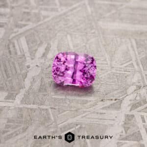 A vibrant 1.24-carat pink Madagascar sapphire with a rectangular cushion cut is placed on a textured, metallic background. The stone sparkles with light reflections, and beneath it is the text "EARTH'S TREASURY" accompanied by a geometric logo.