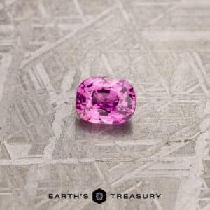 A cushion-cut 1.24-Carat Pink Madagascar Sapphire with a brilliant shine is placed on a textured grey surface. The image has the text "EARTH'S TREASURY" at the bottom along with a geometric logo.