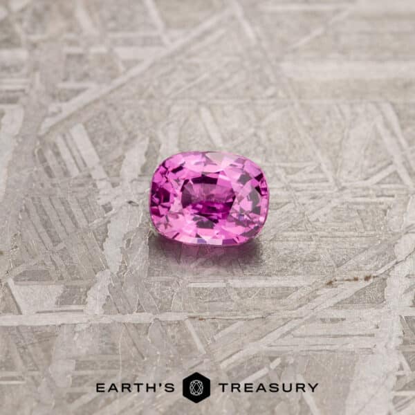 1.01-Carat Hot Pink Madagascar Sapphire A cushion-cut 1.24-Carat Pink Madagascar Sapphire with a brilliant shine is placed on a textured grey surface. The image has the text "EARTH'S TREASURY" at the bottom along with a geometric logo.