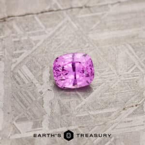 A 1.24-carat pink Madagascar sapphire, expertly cut into a square shape to enhance its sparkle and clarity, is displayed on a textured, angular metallic surface. The text "Earth's Treasury" is visible at the bottom of the image.