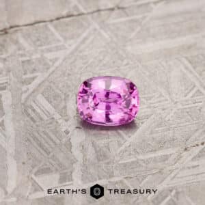 A 1.24-carat cushion-cut pink Madagascar sapphire displayed on a textured metallic surface. The bottom of the image features the logo and text "Earth's Treasury.