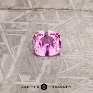 A 1.24-carat pink Madagascar sapphire, cushion-cut and faceted, displayed on a textured gray surface with the brand name "EARTH'S TREASURY" at the bottom.