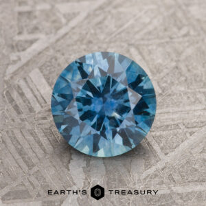 A 1.34-carat teal blue Montana sapphire (heated), round brilliant-cut, is displayed on a textured metallic surface with "EARTH'S TREASURY" at the bottom.