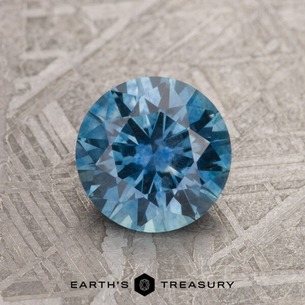 1.34-Carat Teal Blue Montana Sapphire (Heated) A 1.34-carat teal blue Montana sapphire (heated), round brilliant-cut, is displayed on a textured metallic surface with "EARTH'S TREASURY" at the bottom.