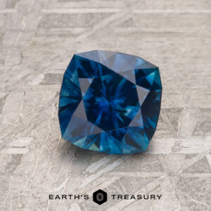 A 1.26-carat royal blue heated Montana sapphire gemstone rests on a textured gray surface with "EARTH'S TREASURY" displayed at the bottom.