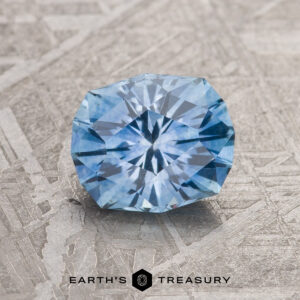 A 1.37-carat Sky Blue Montana Sapphire (heated), round brilliant-cut, is displayed on a textured gray surface with "EARTH'S TREASURY" and a logo at the bottom.