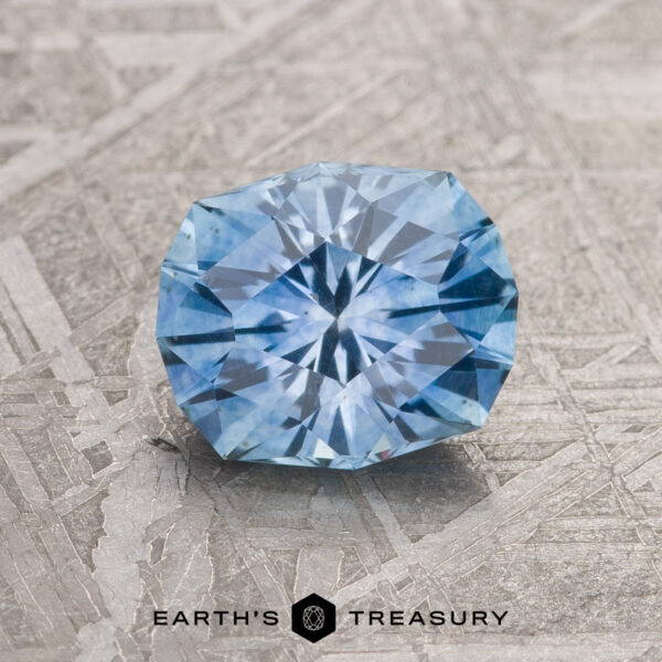 1.37-Carat Sky Blue Montana Sapphire (Heated) A 1.37-carat Sky Blue Montana Sapphire (heated), round brilliant-cut, is displayed on a textured gray surface with "EARTH'S TREASURY" and a logo at the bottom.