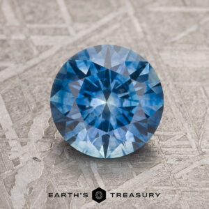 A 1.86-carat medium blue Montana Sapphire (heated), round and faceted, rests on a textured light surface featuring the text "EARTH'S TREASURY" at the bottom.