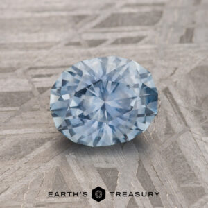A 1.23-Carat Sky Blue Montana Sapphire is shown on a gray patterned surface with "EARTH'S TREASURY" at the bottom.