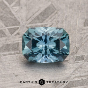 The 1.23-Carat Teal Blue Montana Sapphire, with brilliant facets, is shown on a textured metallic background with "EARTH'S TREASURY" displayed beneath.