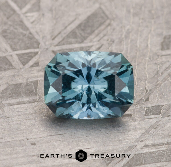 1.23-Carat Teal Blue Montana Sapphire The 1.23-Carat Teal Blue Montana Sapphire, with brilliant facets, is shown on a textured metallic background with "EARTH'S TREASURY" displayed beneath.