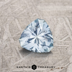A 1.02-carat light sky blue Montana Sapphire, trillion-cut, sits on a textured gray surface above the "EARTH'S TREASURY" logo and text.