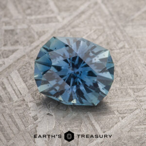 A 1.54-Carat Medium Blue Montana Sapphire with a starburst pattern rests on a textured metallic surface, with the "Earth's Treasury" logo and text at the bottom.