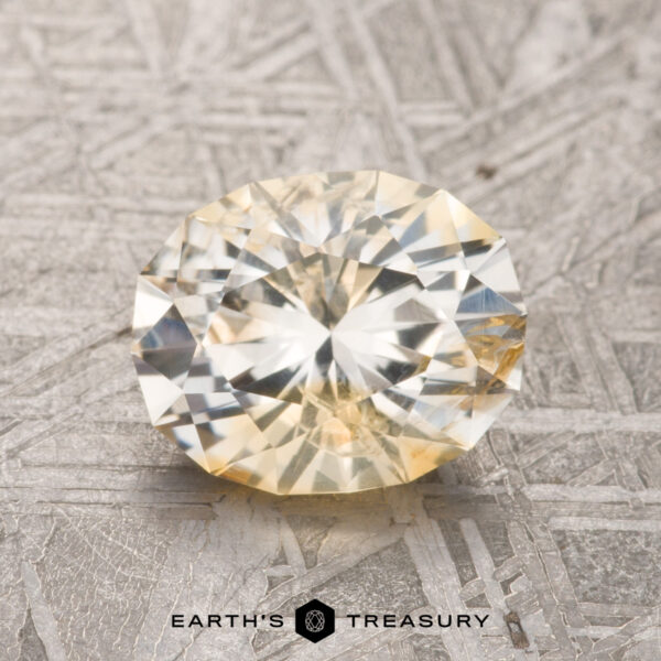 1.93-Carat Colorless-Yellow Particolored Montana Sapphire (Heate A 1.93-carat, faceted round yellow sapphire from Montana sits delicately on a textured gray surface, accompanied by the brand name "Earth's Treasury" at the bottom.