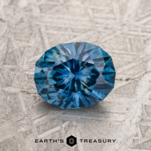 A 1.36-carat medium blue heated Montana Sapphire is shown on a textured silver-gray surface with "EARTH'S TREASURY" at the bottom.