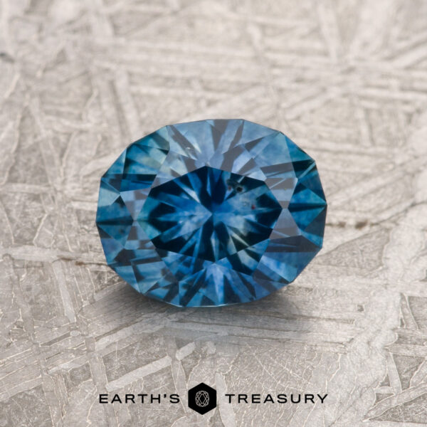 1.36-Carat Medium Blue Montana Sapphire (Heated) A 1.36-carat medium blue heated Montana Sapphire is shown on a textured silver-gray surface with "EARTH'S TREASURY" at the bottom.